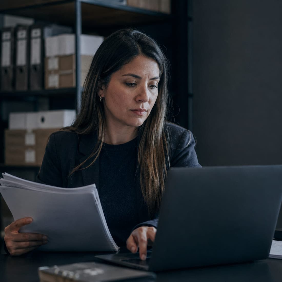Encargada de personas revisando un fajo de contratos de trabajo junto a su computador, con cajas de documentación al fondo